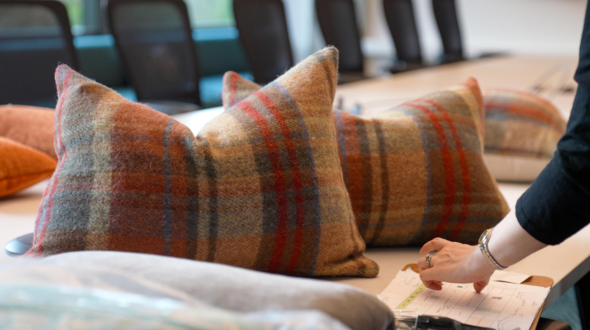 Plaid cushion covers in rust, olive and slate arranged on a conference table, with a hand pointing to a floor plan on a clipboard